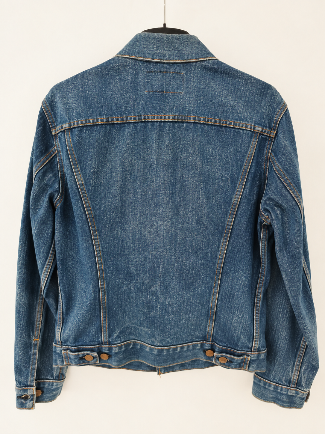 A blue denim jacket shown from the back, hanging on a black hanger against a plain off-white background.
