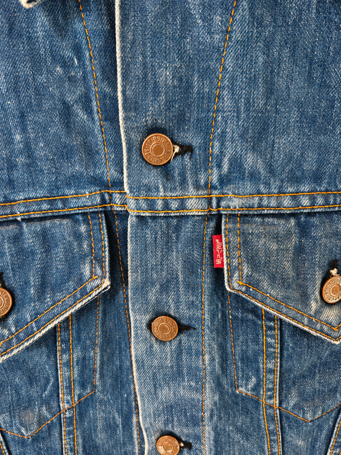 Close-up of a blue denim jacket showing the front button closure with bronze-colored buttons, dual flap pockets, and a small red Levi's brand tab.