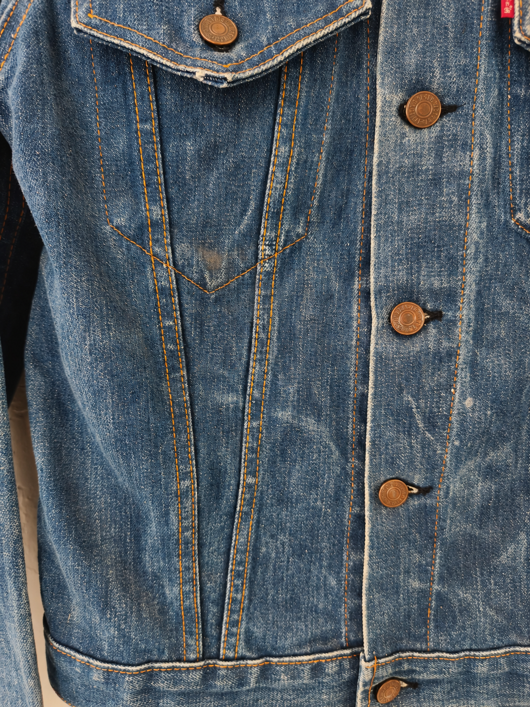Close-up of a blue denim jacket showing the front button closure with bronze-colored buttons, chest pockets, and detailed stitching.