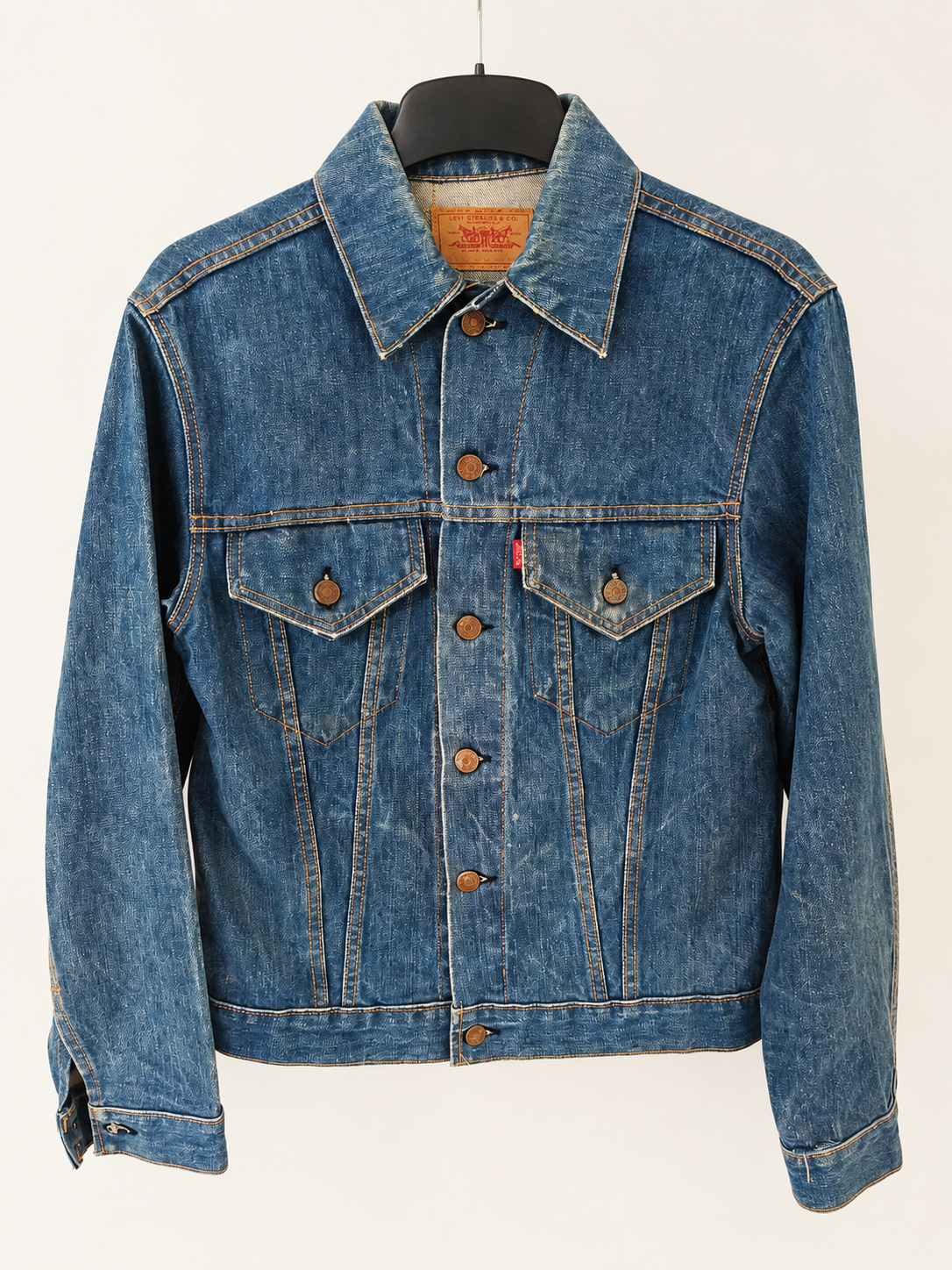 A classic blue denim trucker jacket from Levi's big E hanging on a black hanger against a plain white background. The jacket features a pointed collar, button-up front, two buttoned chest pockets, and signature contrast stitching.