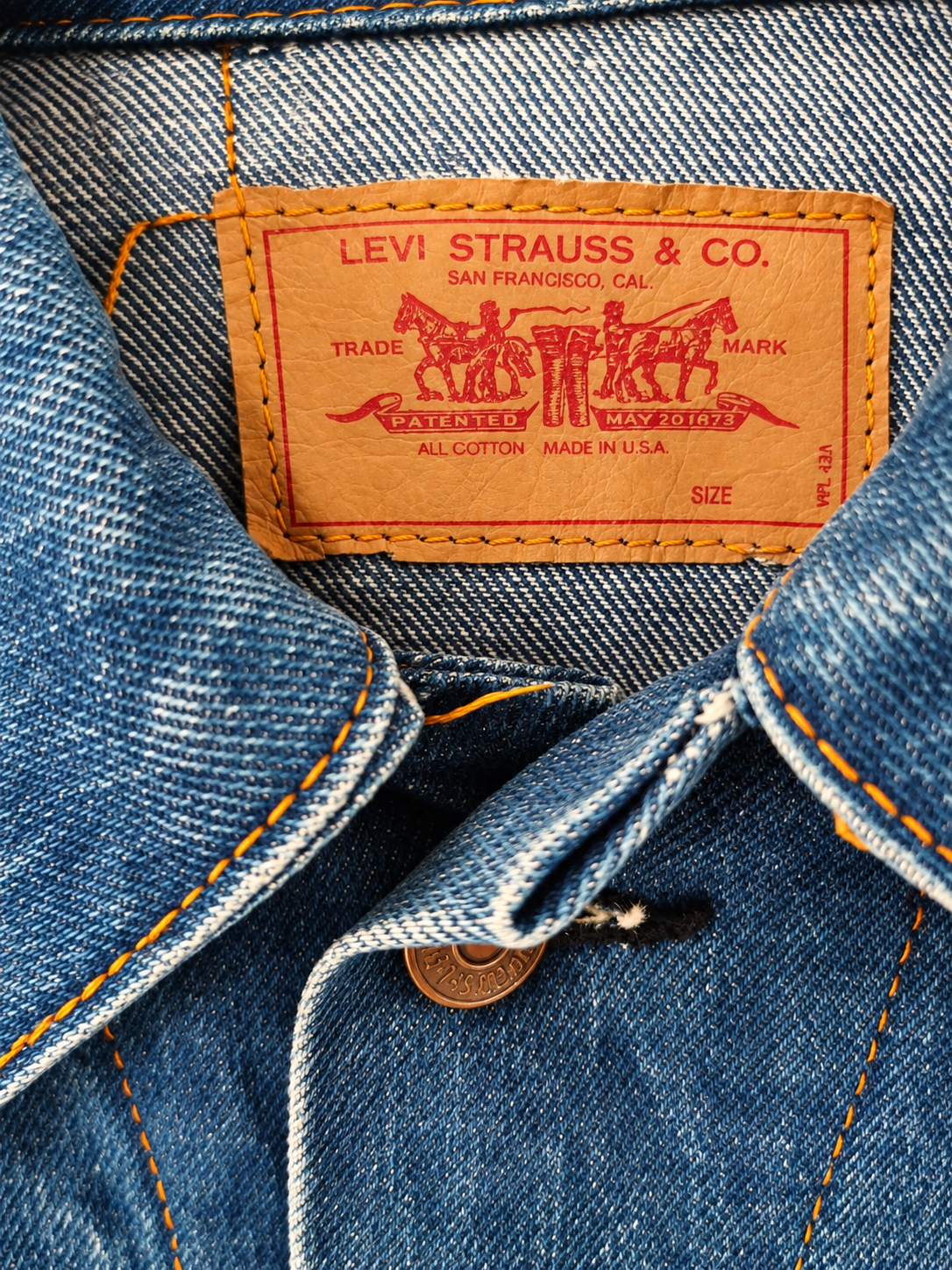 Close-up of a blue denim jacket showing the interior collar with the iconic orange Levi's leather patch and orange contrast stitching.