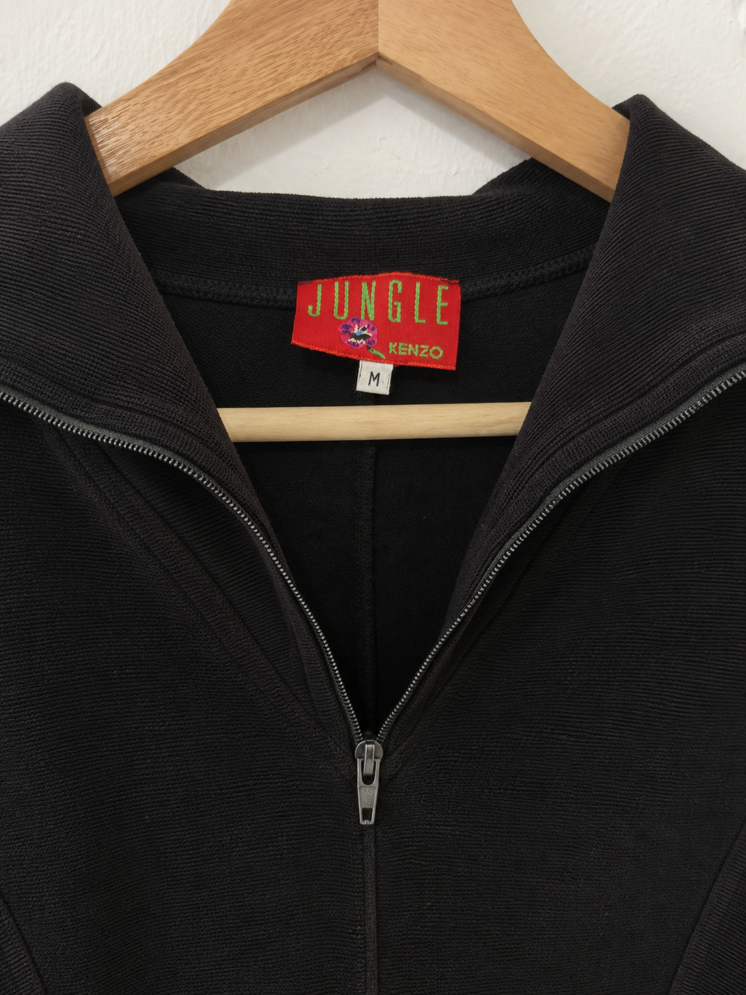 A close-up of a black ribbed knit quarter-zip top hanging on a wooden hanger against a white wall. The red and green Jungle Kenzo brand label is visible inside the collar.