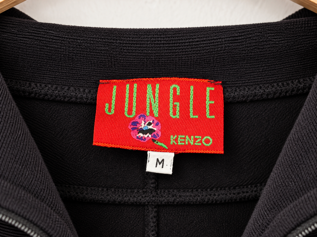 Close-up of a black knit garment showing a red woven brand label that reads JUNGLE KENZO with a small floral logo, plus a small white size tag marked M.